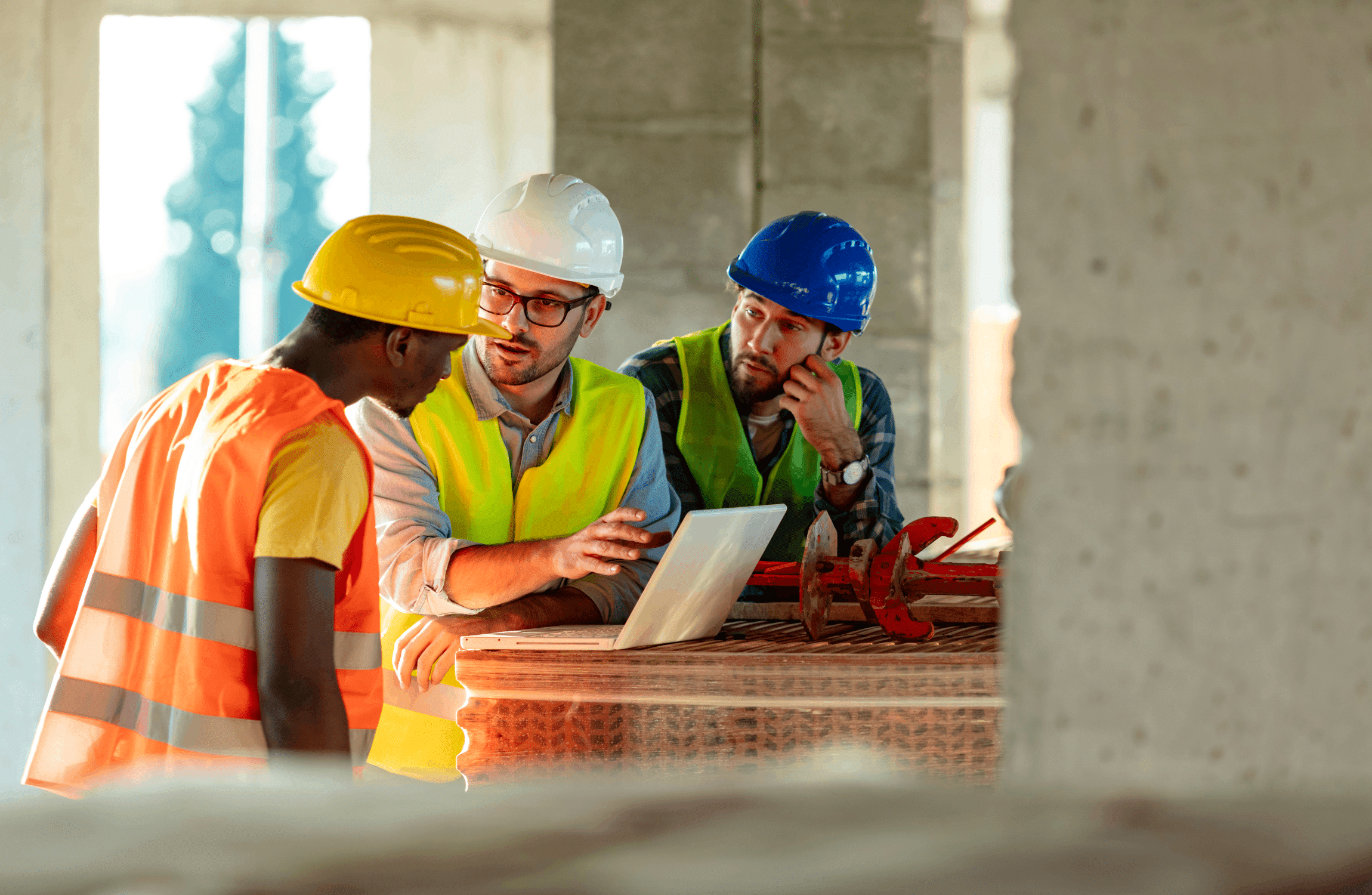 Three construction workers in hard hats and safety vests discussing a project over a laptop inside a building under construction