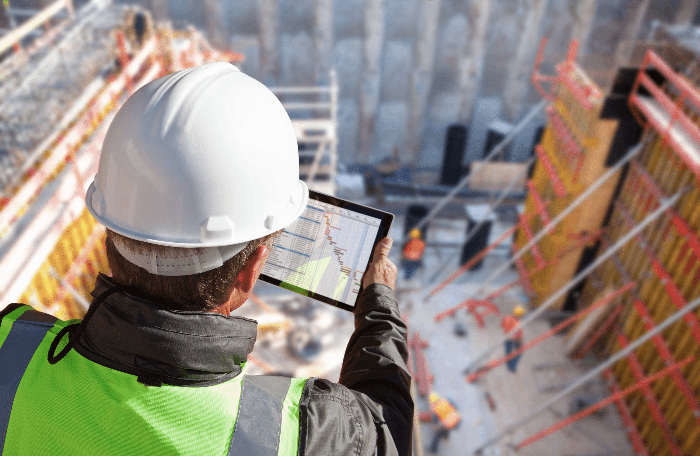 Over-the-shoulder view of a construction manager in a white hard hat and safety vest using a tablet to review a project schedule at a busy construction site