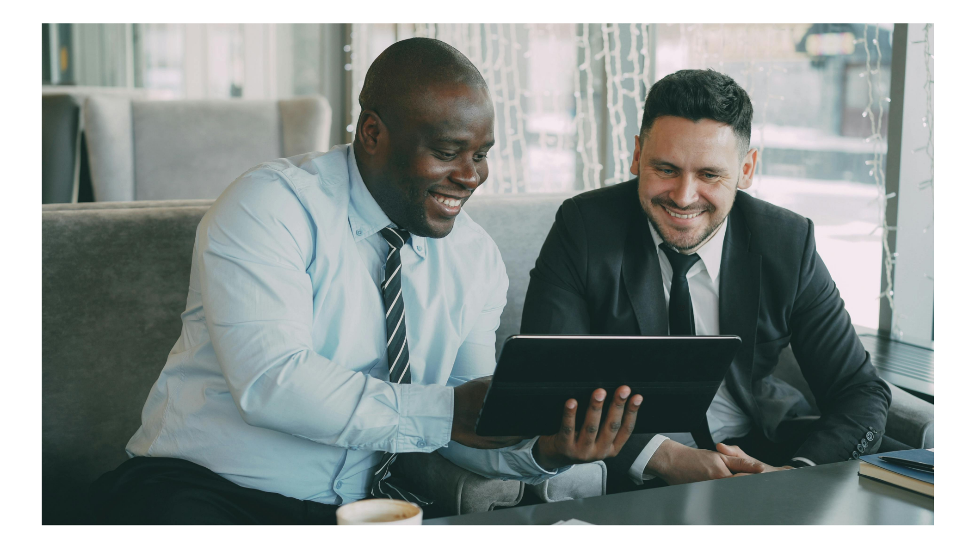 Two smiling businessmen in suits sitting on a sofa and looking at a tablet screen together during a meeting.