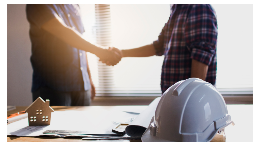 Strategic Construction Partnership and Handshake A close-up view of two people shaking hands in a bright office. In the foreground, a white hard hat, architectural blueprints, and a small wooden house model sit on a wooden desk. The background is softly blurred with natural light coming through a window.