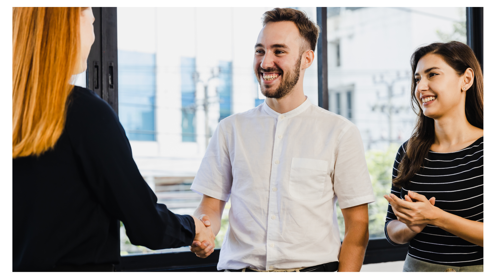 A close-up of a businessman smiling and shaking hands with a woman while a colleague claps in approval by a bright window.