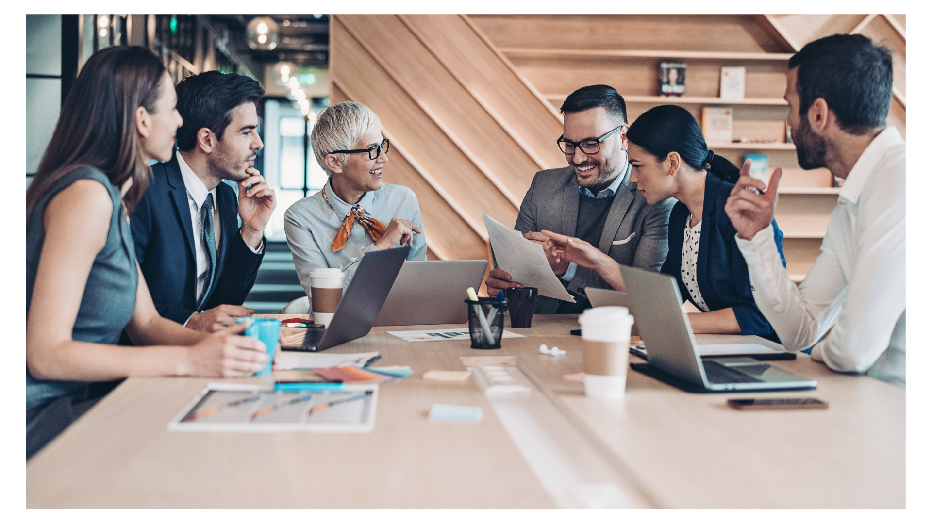 A group of six diverse business executives smiling and collaborating around a conference table with laptops and documents.